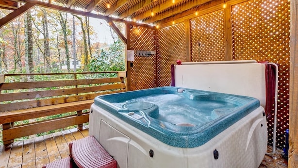A hot tub with a wooden deck and a bench, surrounded by a wooden fence and string lights.