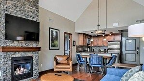 A modern living room with a stone fireplace, a flat-screen TV, and a dining area with blue chairs. - Peaceful Penthouse – Sunrise Views & Resort Amenities! (Canmore)