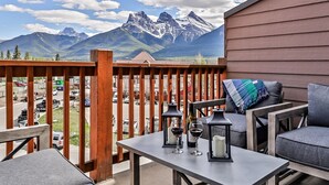 A balcony with a view of mountains, a table with two wine glasses, and a bench with a cushion. - Peaceful Penthouse – Sunrise Views & Resort Amenities! (Canmore)