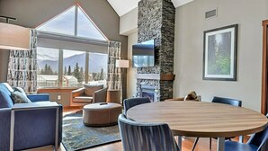 A modern living room with a large window, a stone fireplace, and a round dining table. - Peaceful Penthouse – Sunrise Views & Resort Amenities! (Canmore)