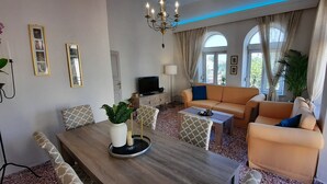 A living room with a wooden dining table, beige sofa, and patterned chairs. There is a TV on a stand, a small side table, and a potted plant. - Philoxenia Accommodation ~ Ralia's House (Κως)