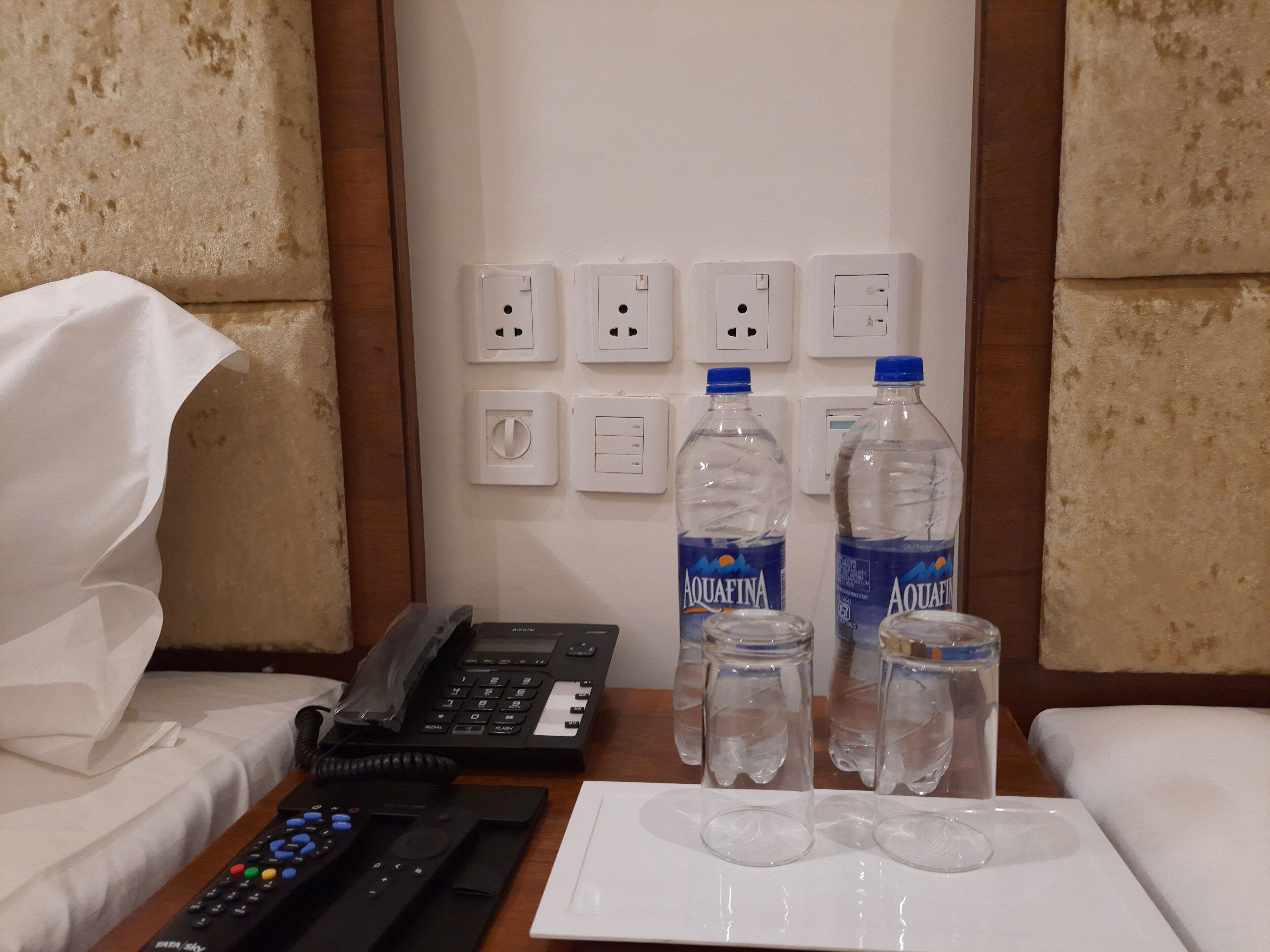 A hotel room with a bed, a bedside table with a phone, remote controls, and two bottles of Aquafina water.