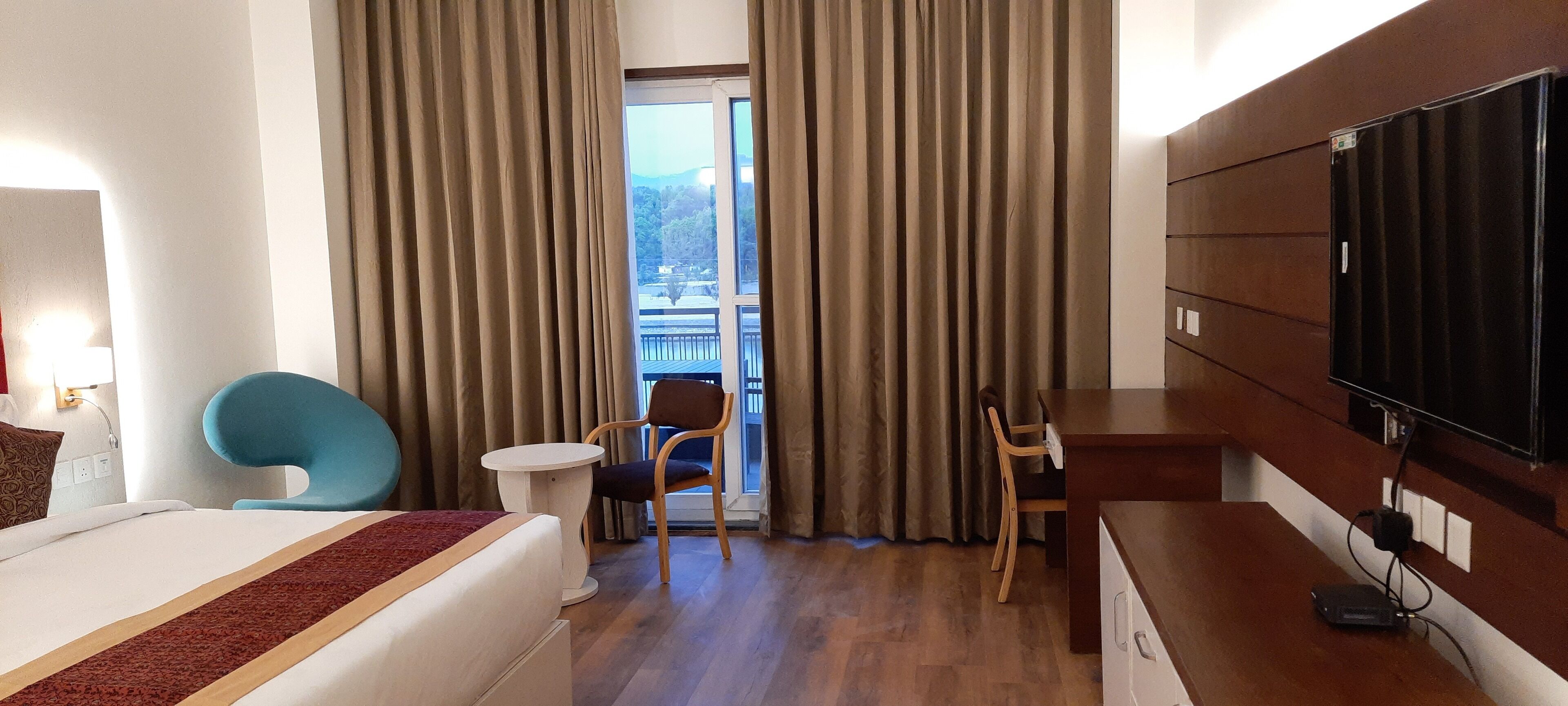 Habitación de hotel con cama, escritorio, silla, televisión y balcón con cortinas.