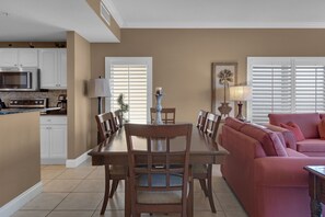 Ein Essbereich mit Holztisch und -stühlen, eine Küche mit weißen Schränken und Geräten sowie ein Wohnzimmer mit rosa Sofa und Lampe.