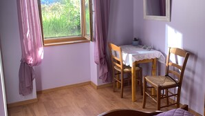 Een slaapkamer met een bed, een raam met gordijnen, een tafel met stoelen en een bijzettafeltje met spullen erop.