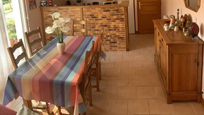 Een eetgedeelte met een tafelkleed met strepen, houten stoelen en een vaas met witte bloemen. Een bakstenen scheidingswand scheidt het eetgedeelte van de keuken.