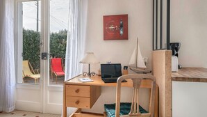 A room with a wooden desk, a laptop, a lamp, a sailboat model, a red chair, and a view of greenery outside. - House avec jardin à 140m de la Plage (Saint-Michel-Chef-Chef)