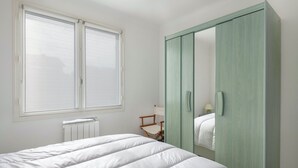A bedroom with a bed, a window with blinds, a radiator, a wooden chair, and a green wardrobe. - House avec jardin à 140m de la Plage (Saint-Michel-Chef-Chef)