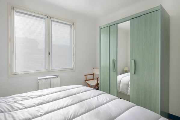 Un dormitorio con cama, ventana con persianas, radiador, una silla de madera y un armario verde.