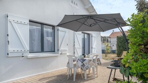A patio with a table and chairs, a large umbrella, and a white building with shutters. - House avec jardin à 140m de la Plage (Saint-Michel-Chef-Chef)