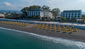 Un hôtel situé en bord de mer, avec des rangées de chaises longues et de parasols, un espace piscine et des bâtiments en arrière-plan.