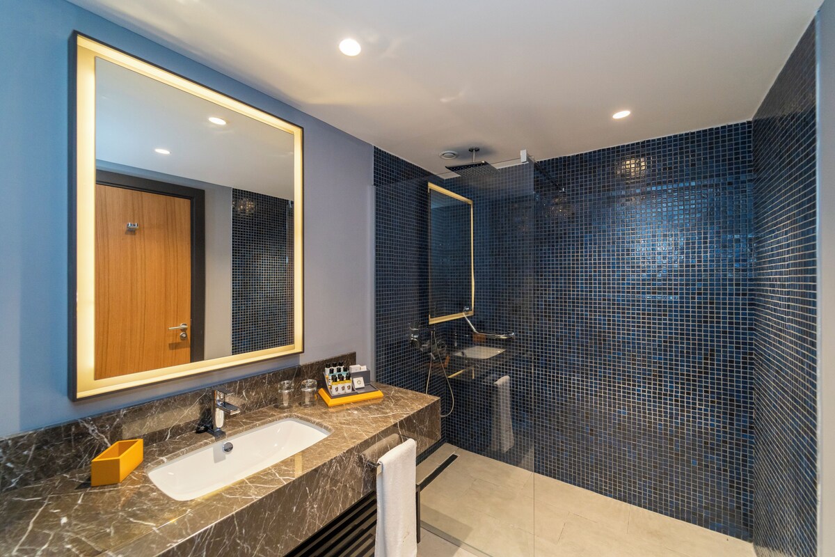 A modern bathroom with a marble sink, a large mirror, and a shower with blue mosaic tiles.