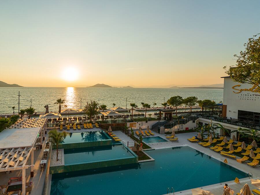 A pool area with multiple sections, sun loungers, and umbrellas, overlooking a body of water with a sunset in the background.