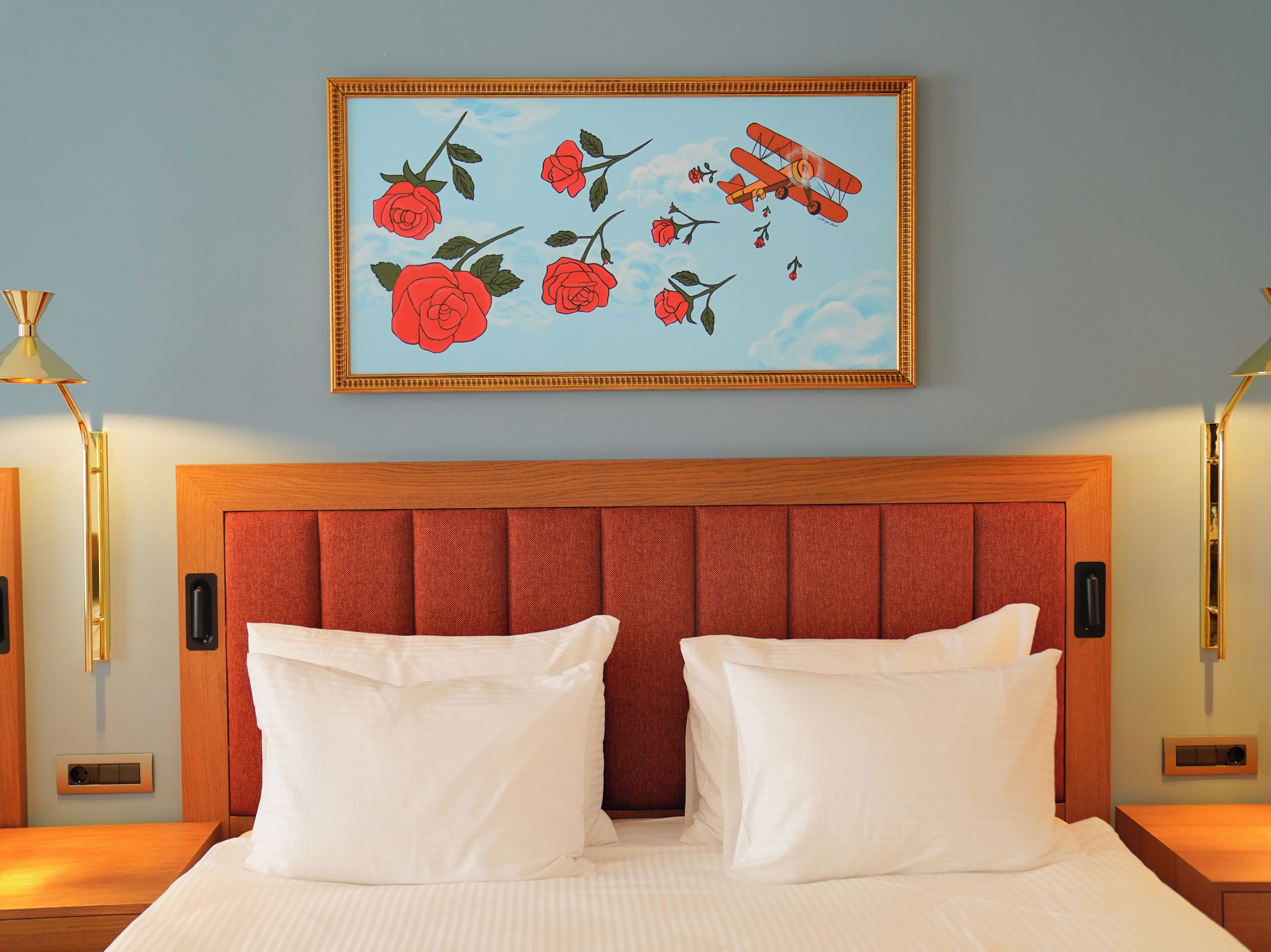 A hotel room with a bed, a headboard, a painting of roses and an airplane, and two bedside lamps.