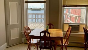 A dining area with a table and chairs, a window with blinds, and a view of a wooden deck and a building. - Charming 3rd Floor Walk-up River View (Clayton)
