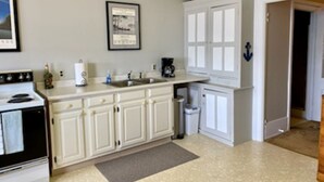 A kitchen with white cabinets, a stove, a sink, and a trash can. There are framed pictures on the wall and a door with an anchor symbol. - Charming 3rd Floor Walk-up River View (Clayton)