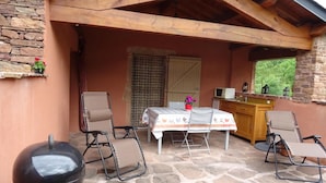 An outdoor patio with stone flooring, a table with a tablecloth, two chairs, a barbecue grill, and a kitchen area with a microwave.