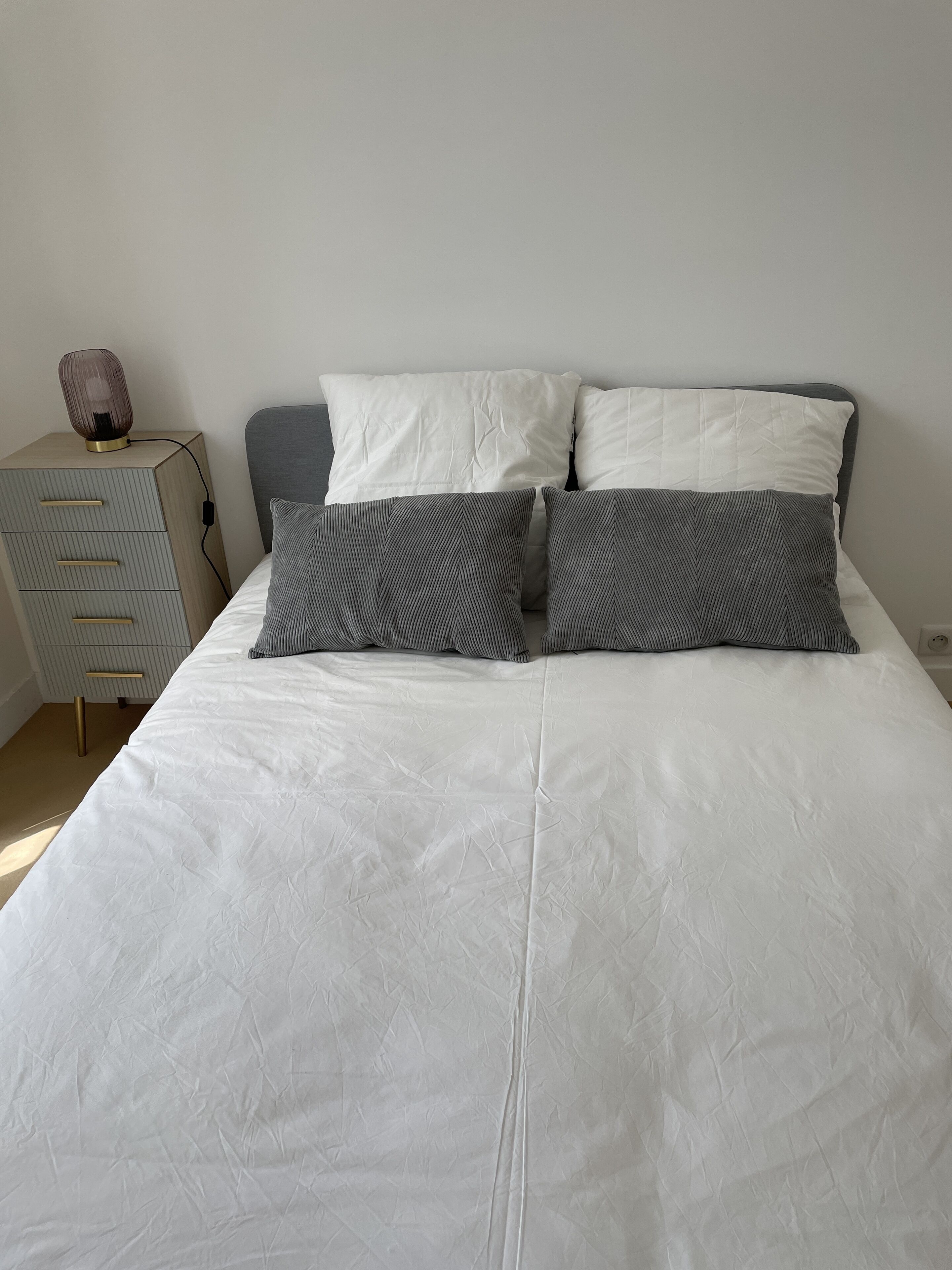 A neatly made bed with a white duvet and two pillows, a bedside table with a lamp, and a drawer unit.