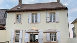 Une maison à deux étages dotée d’un balcon, de volets blancs, ainsi que d’une table et d’un banc en bois installés pour les repas en plein air.