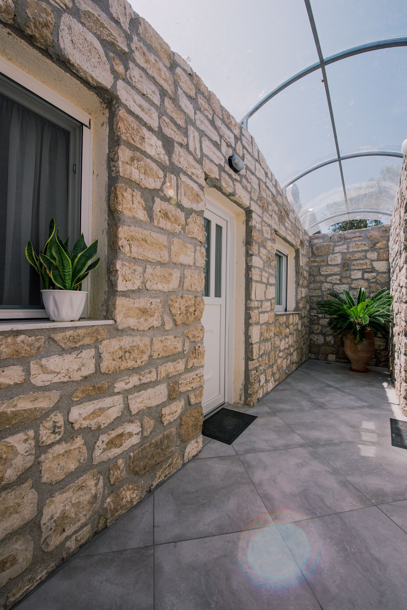 A stone-walled corridor with a glass ceiling, potted plants, and a door.