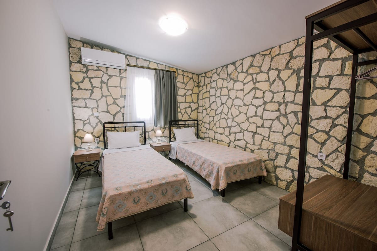 A bedroom with two beds, stone walls, a wooden table, and an air conditioning unit.