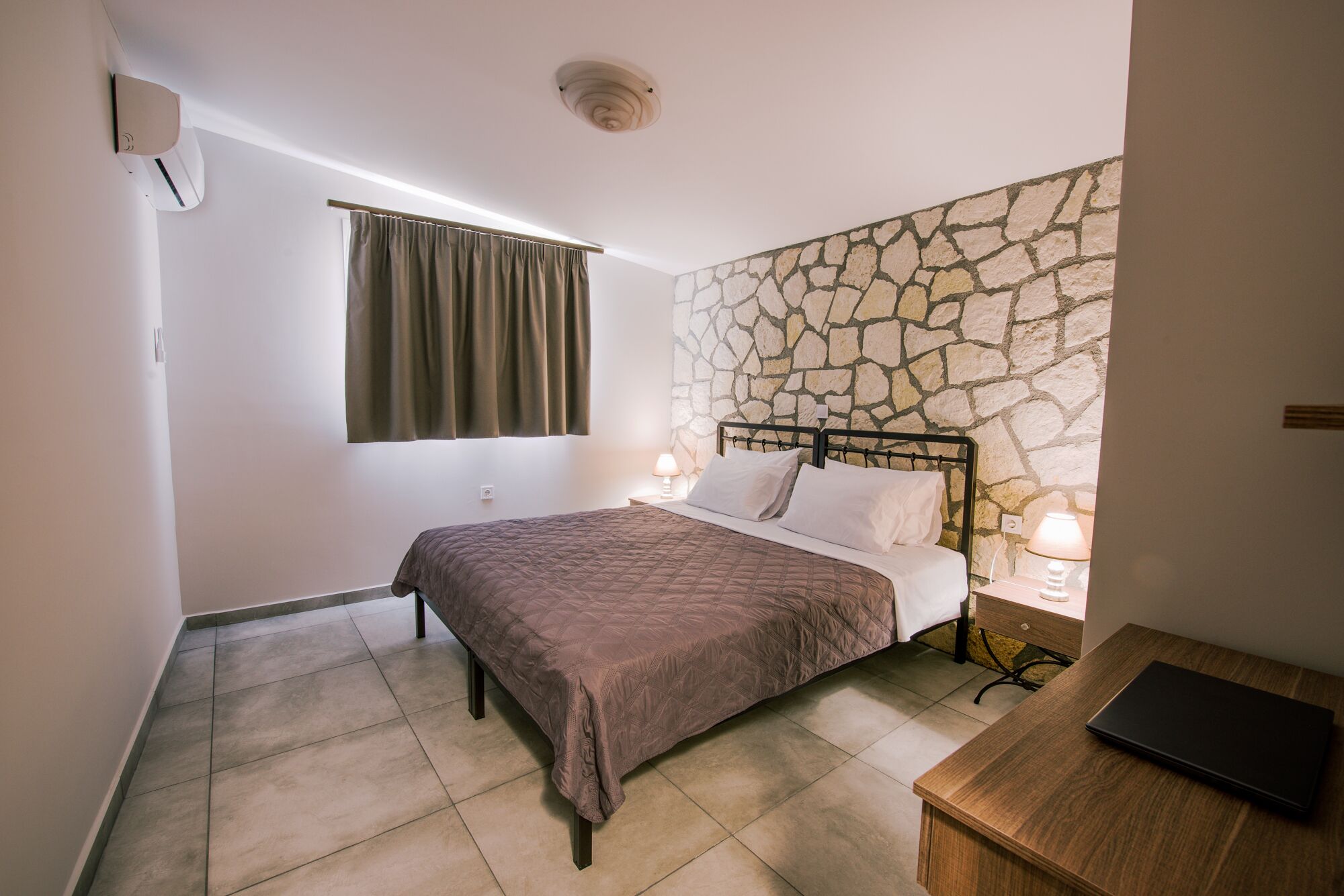 Une chambre avec un mur en pierre, un lit avec une tête de lit, des lampes de chevet et un bureau avec un ordinateur portable.
