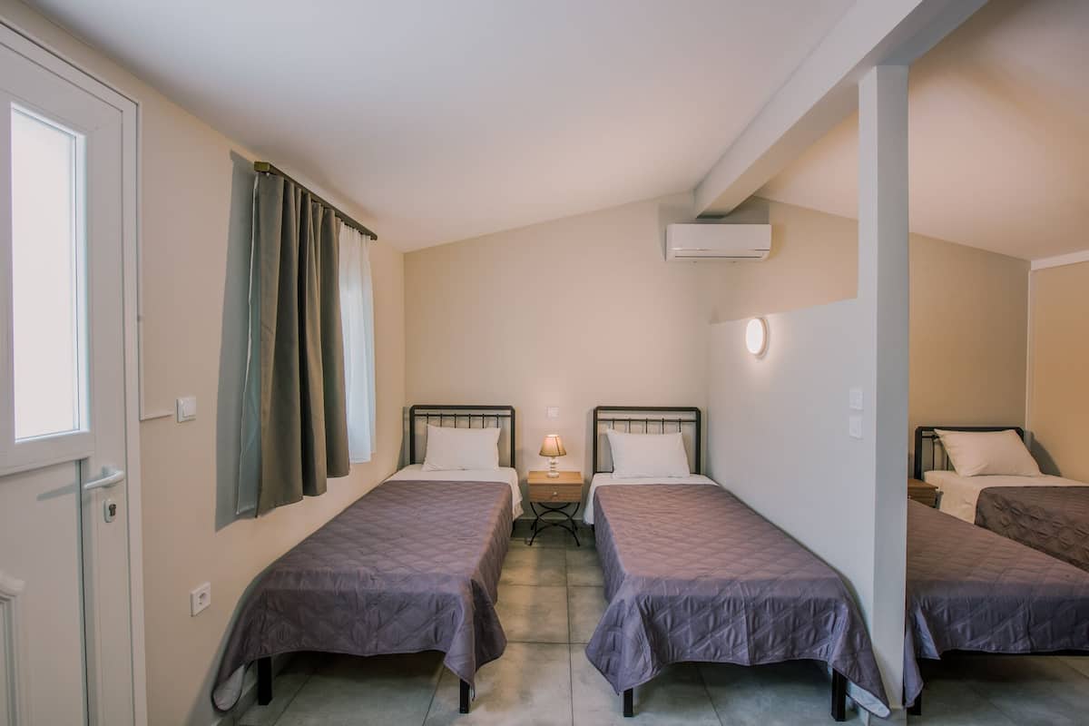 A hotel room with three single beds, a bedside table with a lamp, an air conditioning unit, and a window with curtains.