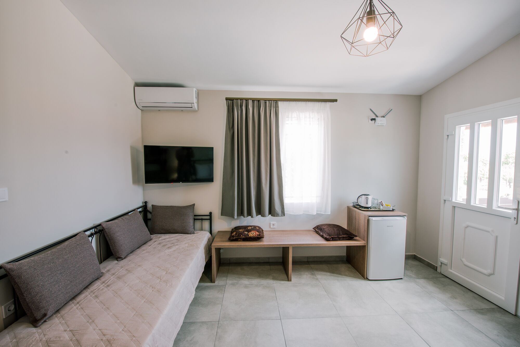 A compact room with a sofa, a small table, a TV, a mini-fridge, and a window with curtains.