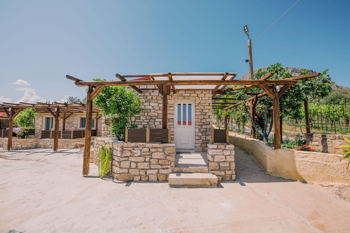 A stone building with a wooden pergola entrance, surrounded by greenery and a clear sky.