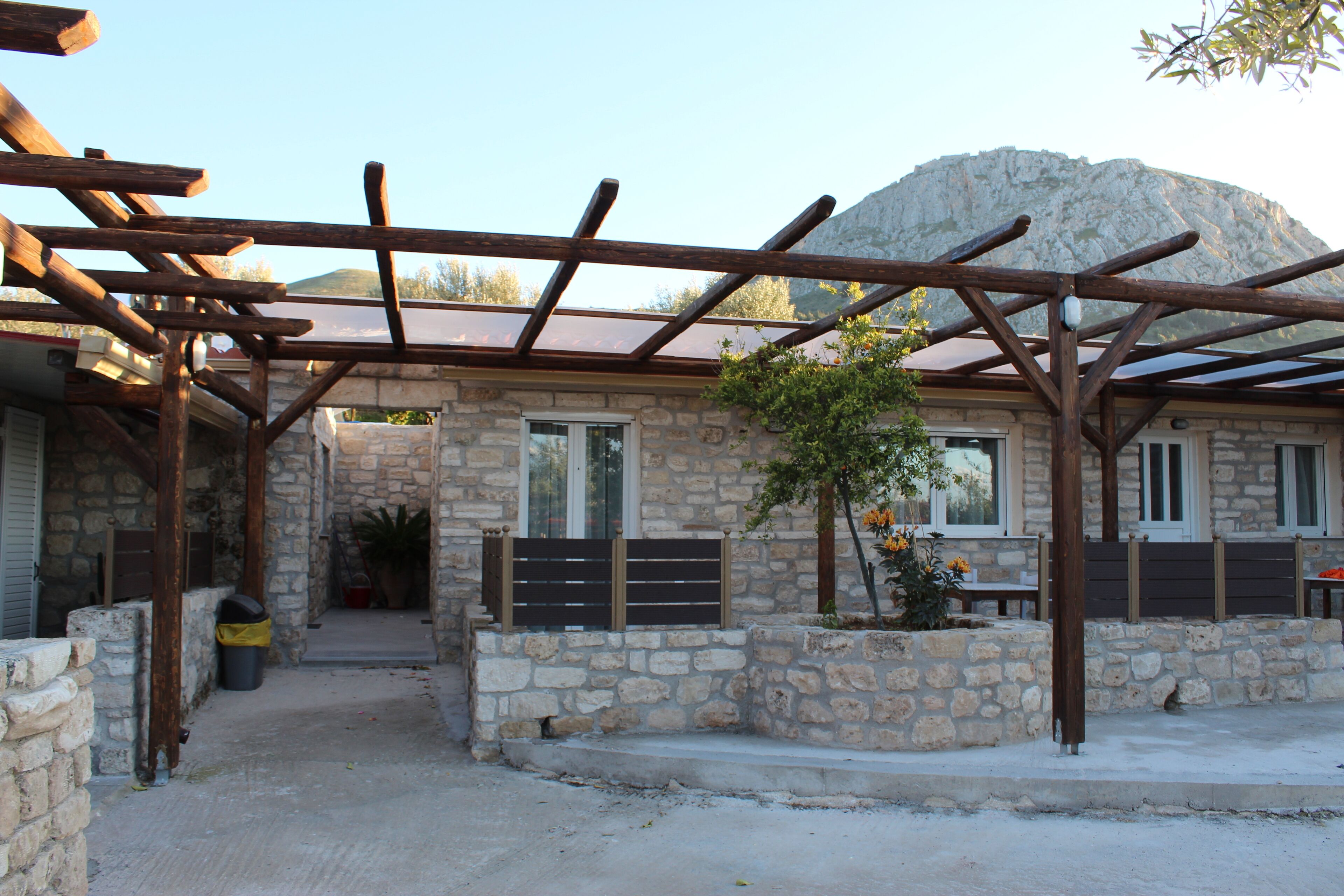 Un edificio de piedra con una pérgola de madera, muros de piedra y una montaña al fondo.
