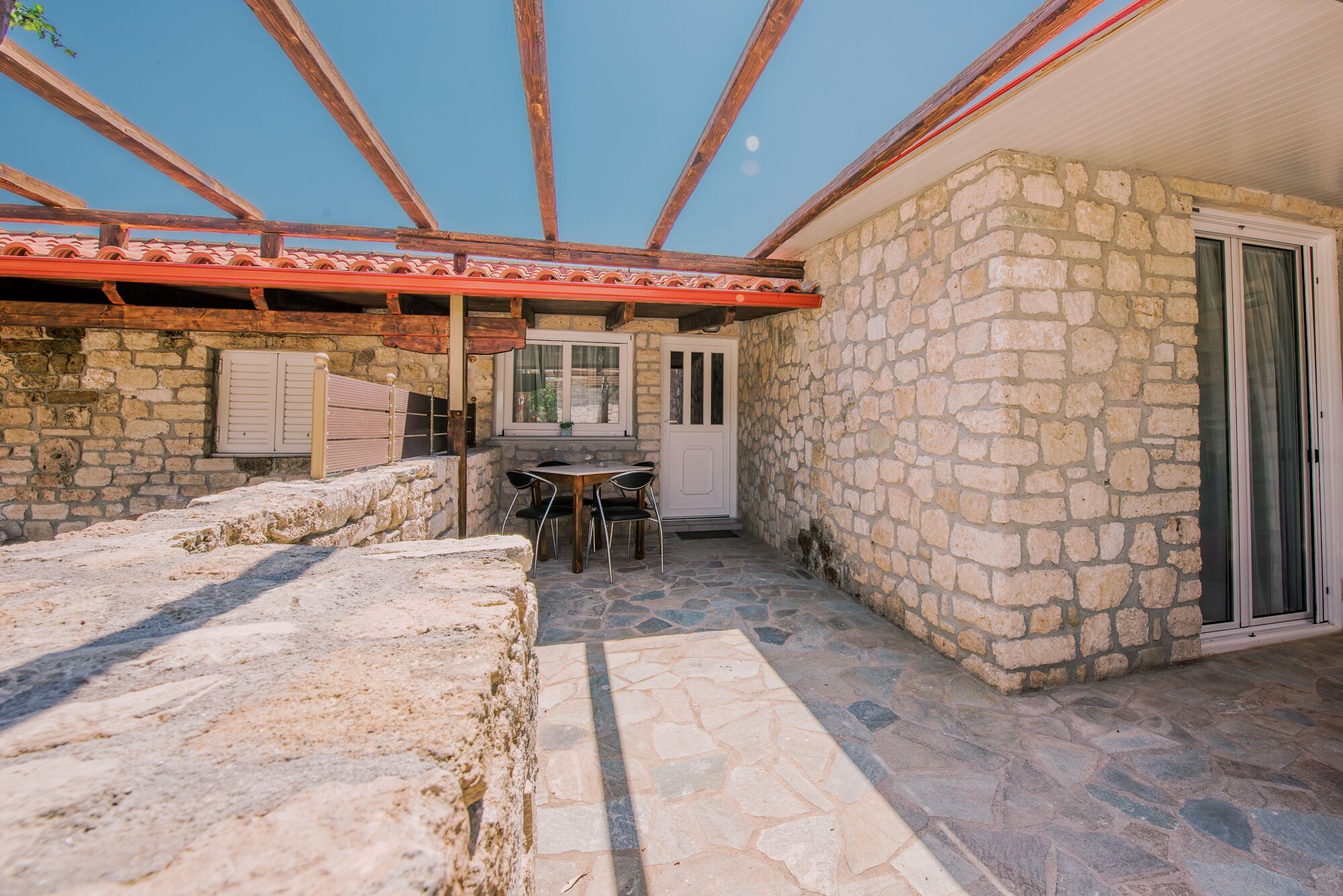 A stone-walled outdoor area with a wooden pergola, a table with chairs, and a door leading to a covered entrance.