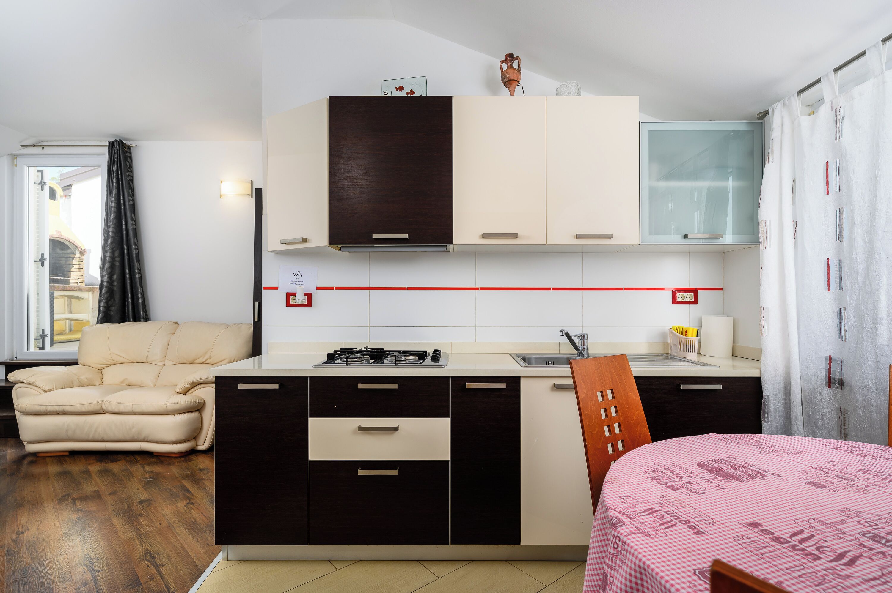 A compact kitchen with a gas stove, a sink, and a small countertop. There is a sofa and a bed with a checkered bedspread.