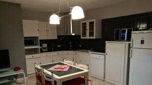 Et kompakt køkken med hvide skabe, et spisebord med stole, en mikrobølgeovn, kaffemaskine og et køleskab.