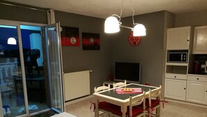 Et spiseområde med et bord og stole, et fjernsyn samt et køkken med skabe og hårde hvidevarer.