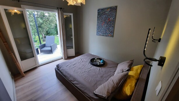 A bedroom with a bed, a bedside table, a chair outside the door, and a painting on the wall.