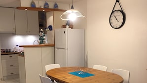Une cuisine compacte avec des meubles blancs, un réfrigérateur blanc et un coin repas comprenant une table en bois et des chaises blanches. Une horloge murale est fixée au mur au-dessus de la table à manger.