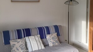 Une pièce avec un canapé gris, des coussins à rayures, un tableau encadré au mur, un lampadaire, une table basse blanche avec un objet bleu dessus, et une porte en bois.