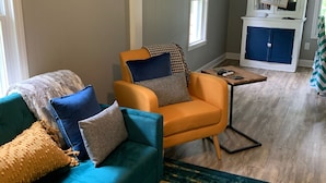 Una sala de estar con un sofá color turquesa, un sillón naranja y otro sillón azul. Hay una mesa de centro de madera y un aparador azul con un letrero encima.