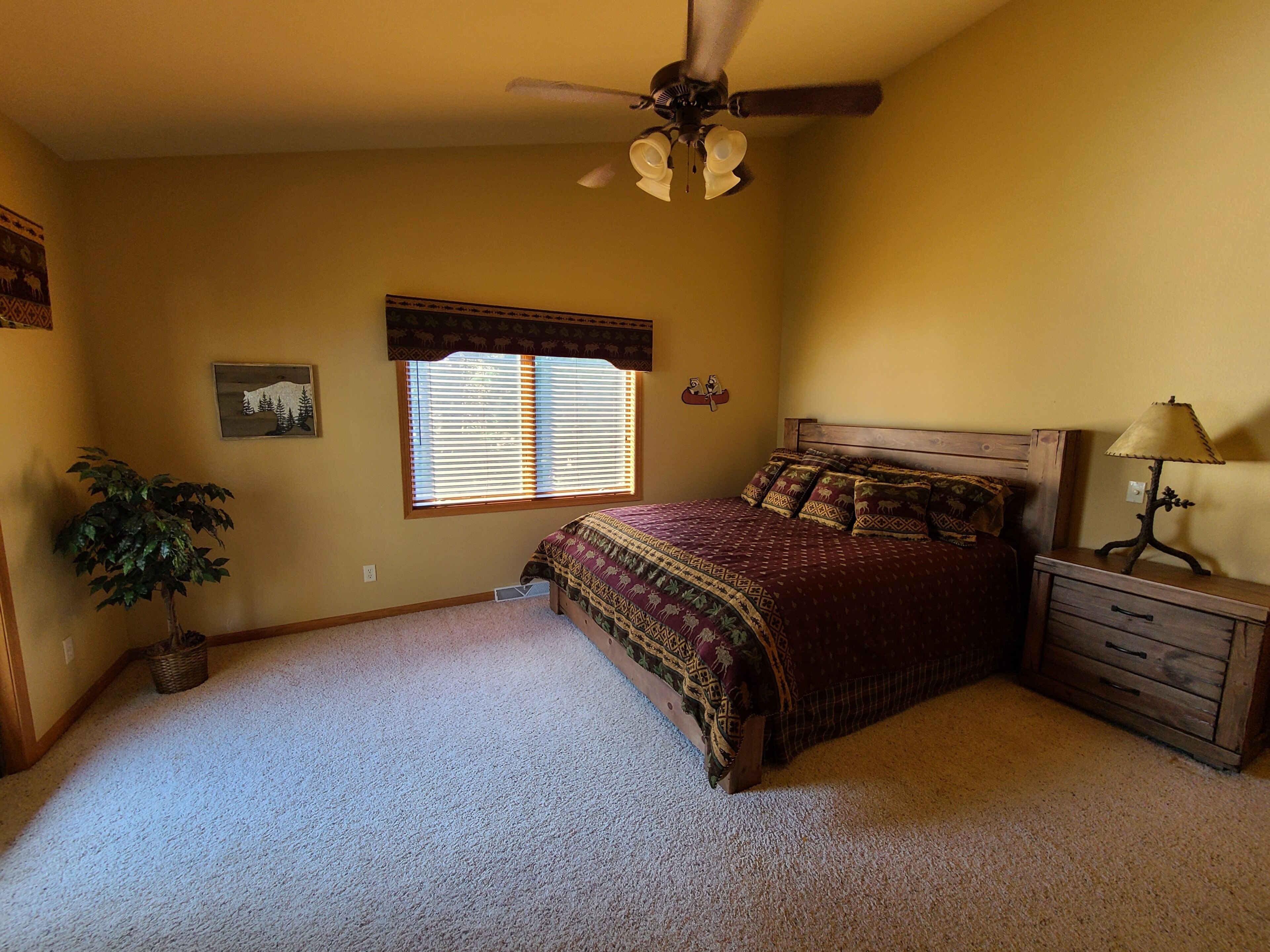 A bedroom with a bed, a nightstand, a lamp, a ceiling fan, a window with blinds, a wall-mounted picture, and a potted plant.