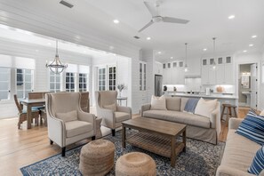 Un vaste espace de vie comprenant un grand canapé, des fauteuils et une table basse. On y trouve également un coin repas avec des chaises et une table en osier. La cuisine, avec ses meubles blancs et son réfrigérateur, est visible au fond.