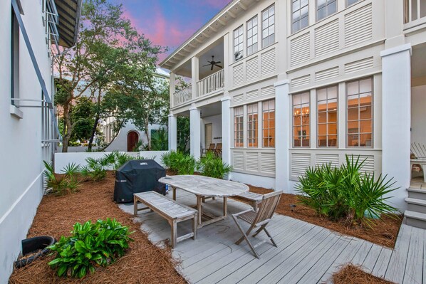 A patio with a wooden table and benches, surrounded by plants and a grill.