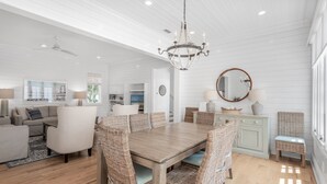 A spacious living room with a large wooden dining table surrounded by wicker chairs, a chandelier, and a view of the kitchen and dining area. - Access to Fitness Center and 4 Pools (Inlet Beach)