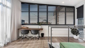 Una habitación moderna con escritorio, dos sillas, un reposapiés verde, una ventana con persianas y una estantería metálica.