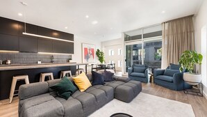 Una sala moderna con un sofá seccional gris, un área de cocina con gabinetes oscuros y un ventanal con cortinas.