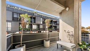 Un balcón moderno al aire libre con una banca, plantas en macetas y vistas de un edificio de varios pisos con balcones.