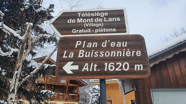 Un panneau indicateur en bois indiquant les directions vers le « télésiège du Mont de Lans (village), accès gratuit aux piétons » et vers le « lac de La Buissonnière, altitude 1 620 m ».