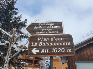 Ein hölzernes Wegweiser mit Hinweisen zum 'Télésiège de Mont de Lans (Village) Gratuit - piétons' und zum 'Plan d'eau La Buissonnière Alt. 1620 m'.