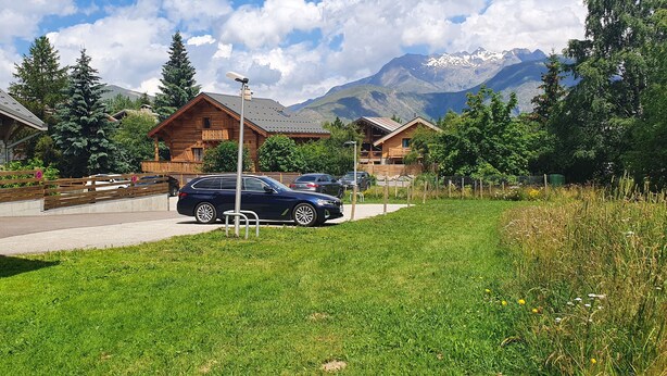 Eine Holzhütte mit einem geparkten Auto davor, umgeben von üppigem Grün und Bergen im Hintergrund.