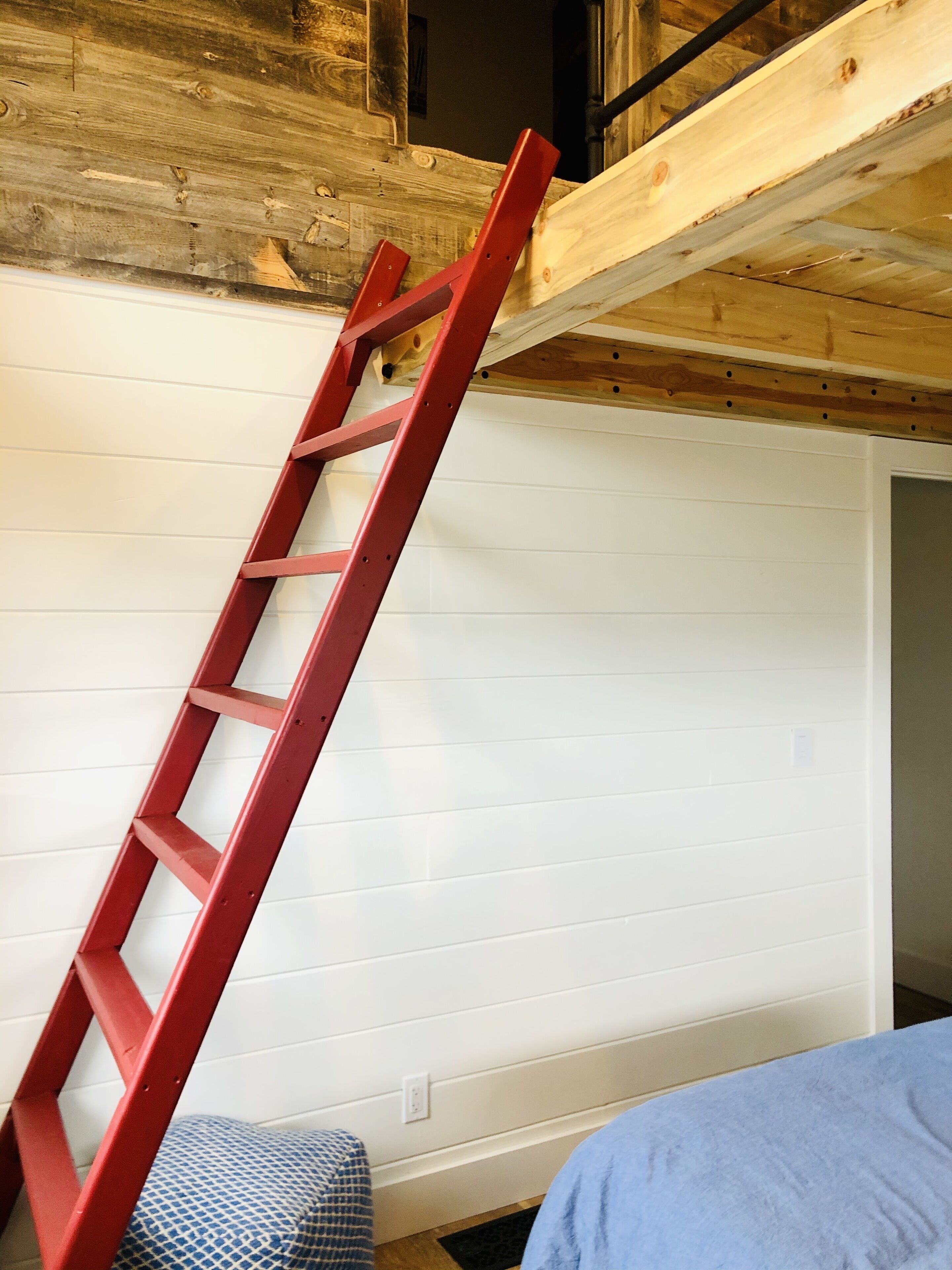 Una stanza con una scala rossa appoggiata a un muro bianco, un soffitto in legno e un letto blu.