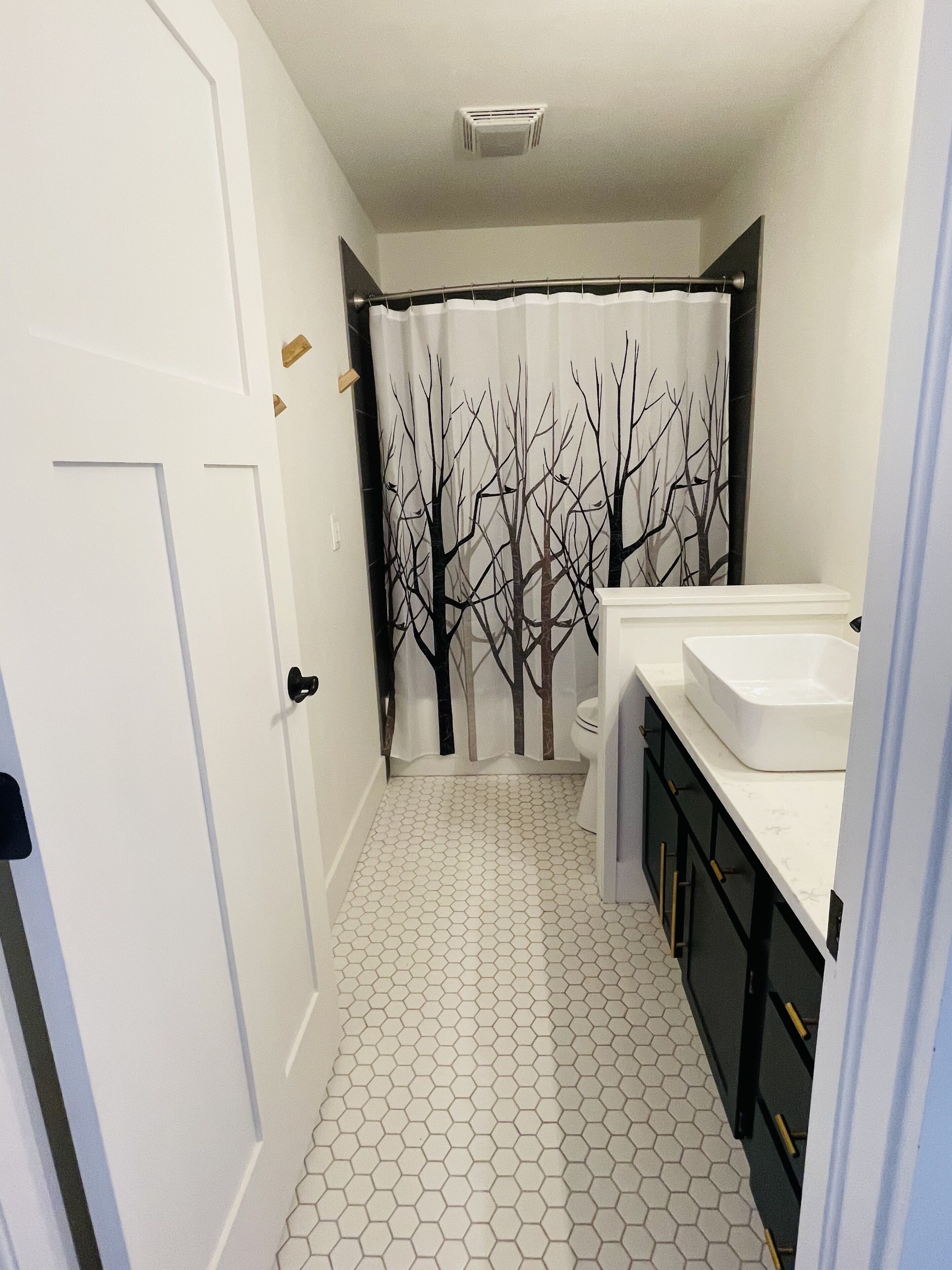 Une salle de bain avec une porte blanche, un rideau de douche orné d’un motif d’arbre, des toilettes et un lavabo avec un plan de travail blanc.